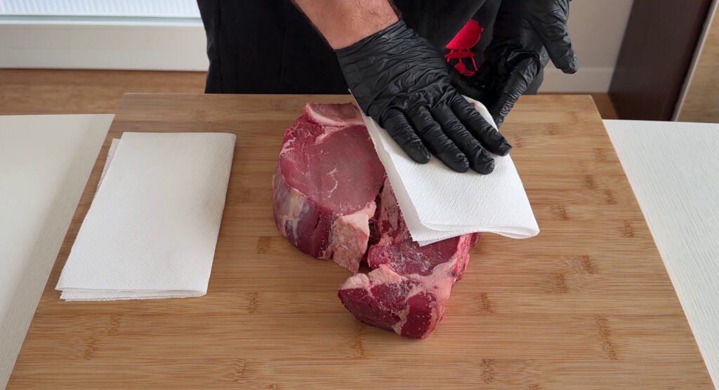 Mani che asciugano una bistecca cruda con carta da cucina, passaggio fondamentale per non far attaccare la carne alla griglia.