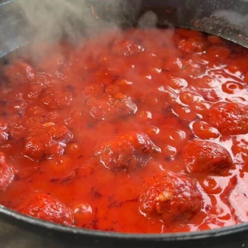 Un classico dutch oven in ghisa posizionato sulle griglie del barbecue, pieno di polpette affumicate che terminano la cottura nel sugo