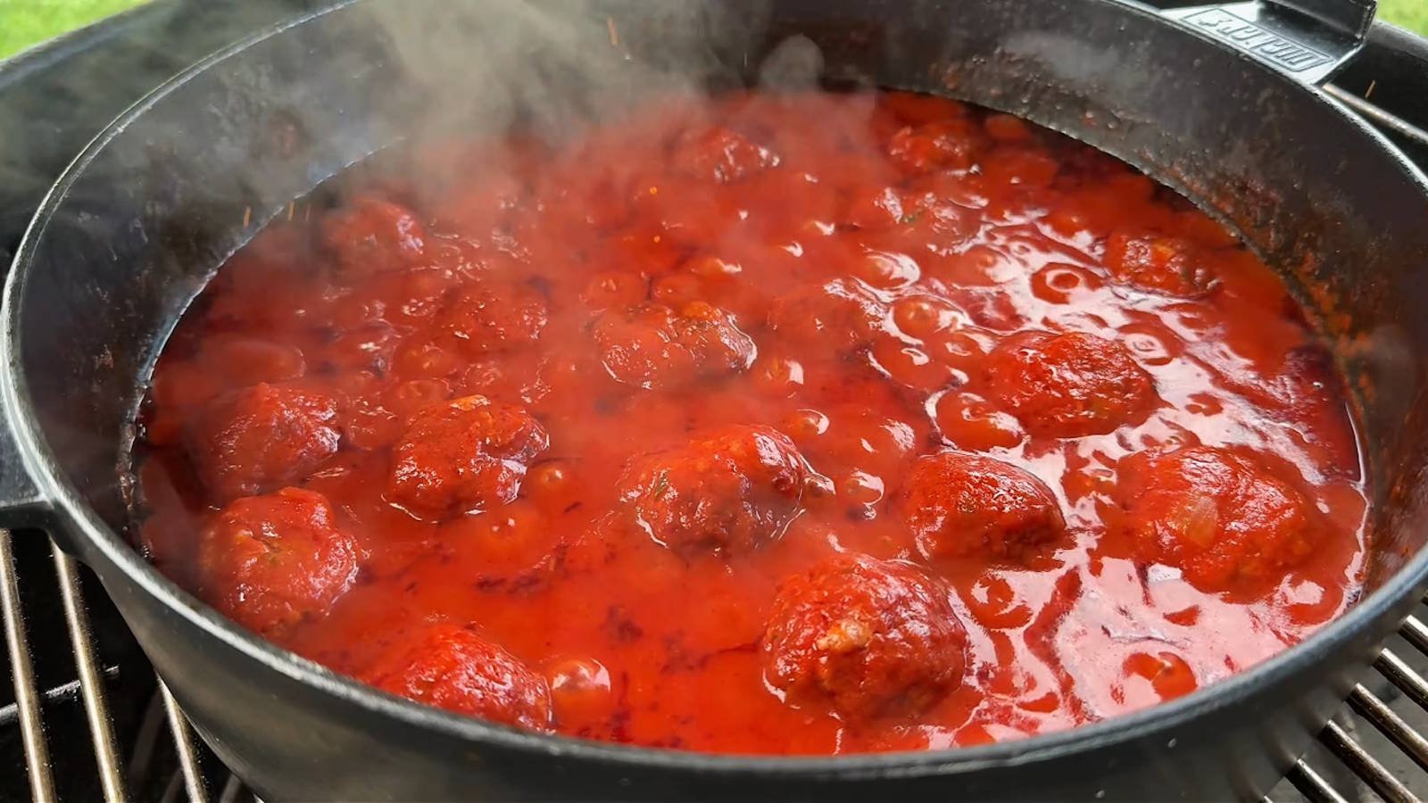 Un classico dutch oven in ghisa posizionato sulle griglie del barbecue, pieno di polpette affumicate che terminano la cottura nel sugo