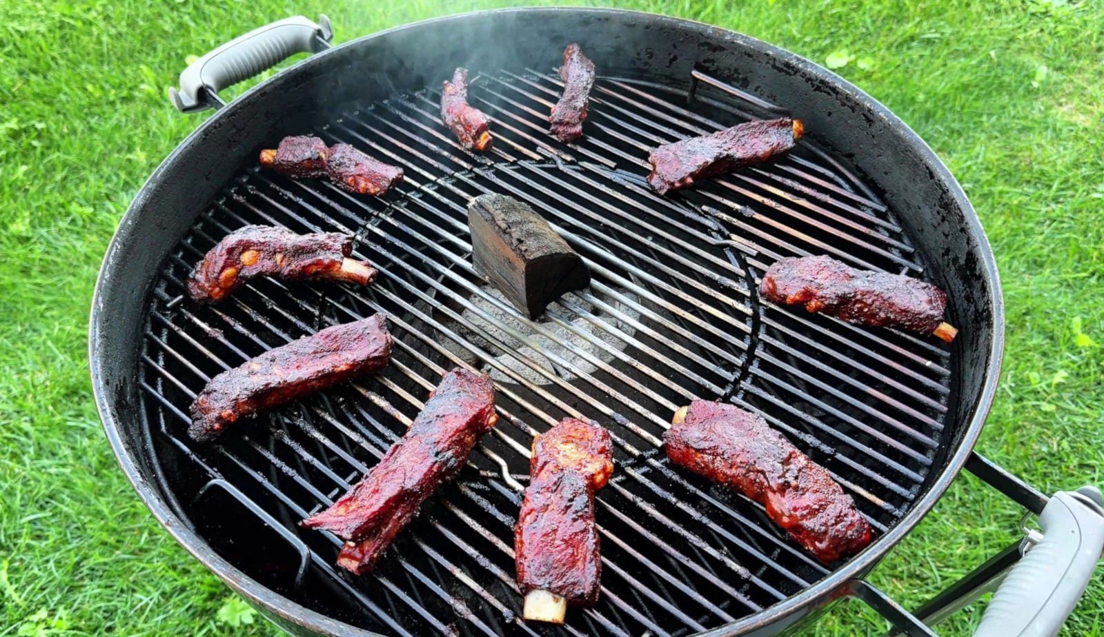 Party ribs glassate disposte ad anello sulla griglia del kettle Weber, intorno al Vortex con un chunk di legno al centro